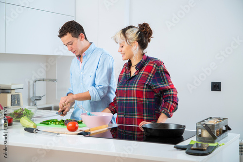 chico joven sonriente corta pimientos verdes con su pareja en una cocina blanca en la que hay mas verduras como tomate, calabacin y lechuga, utilizan una tabla de cortar, un bol rosa y una sarten
