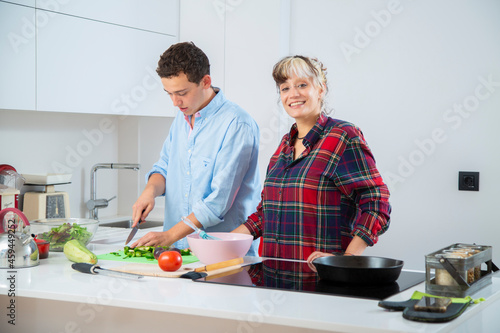 pareja joven sonriente cocinando verduras pimiento verde, tomate y calabacin, en una cocina blanca con placa de induccion, utilizan un bol rosa, una sarten de hierro y usan herramientas de madera