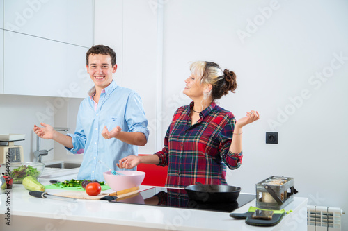 pareja joven sonriente cocina pimiento verde con una sarten de hierro en una cocina blanca con verduras, con placa de induccion, utilizan un bol rosa y usan herramientas de cocina
