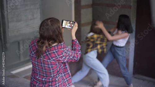 Back view teenage girl filming fight of two angry classmates on smartphone. Caucasian adolescent teenagers fighting outdoors arguing as groupmate recording video for social media photo