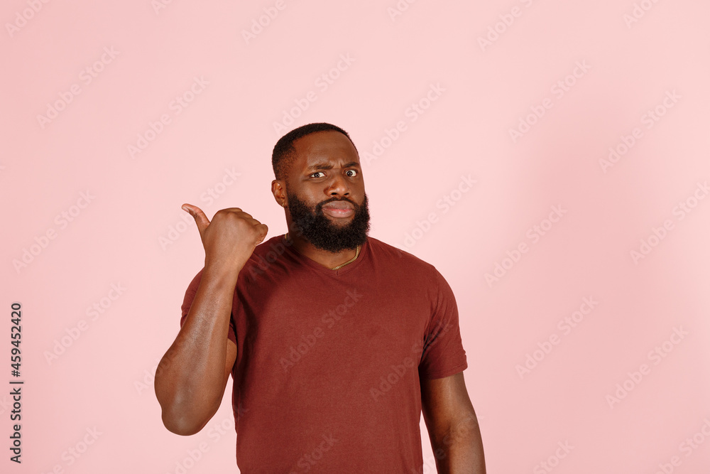 Doubting bearded African-American man in stylish brown t-shirt points aside standing on pink background in studio close view Doubting bearded African-American man in stylish brown t-shirt points aside standing on pink background in studio close view