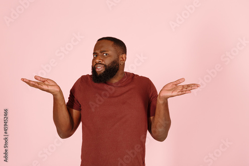 Handsome confused African-American model in stylish brown t-shirt shrugs standing on light pink background in studio closeup