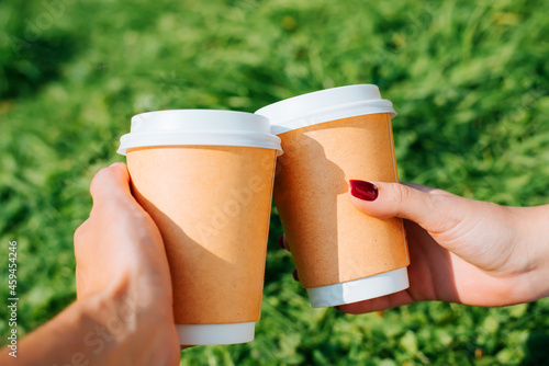 Obraz na plátně Close-up of hand holding two disposable cups of coffee, woman's and man's hand clink glasses outdoors on green lawn
