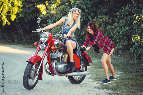 Young beautiful women ride a retro motorcycle. A talented girl in a blue overalls. They push the motorcycle  the merry go to the road. sale of spare parts. Place for text