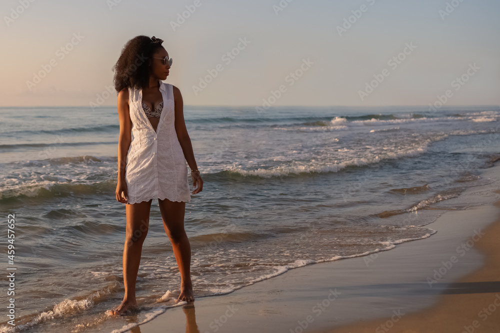 Young adult woman with sunglasses standing along the beach at sunset