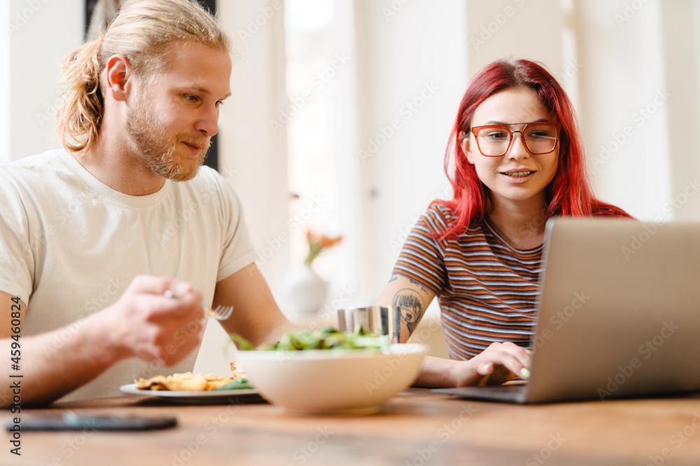 Young white couple using laptop and having lunch while sitting at table