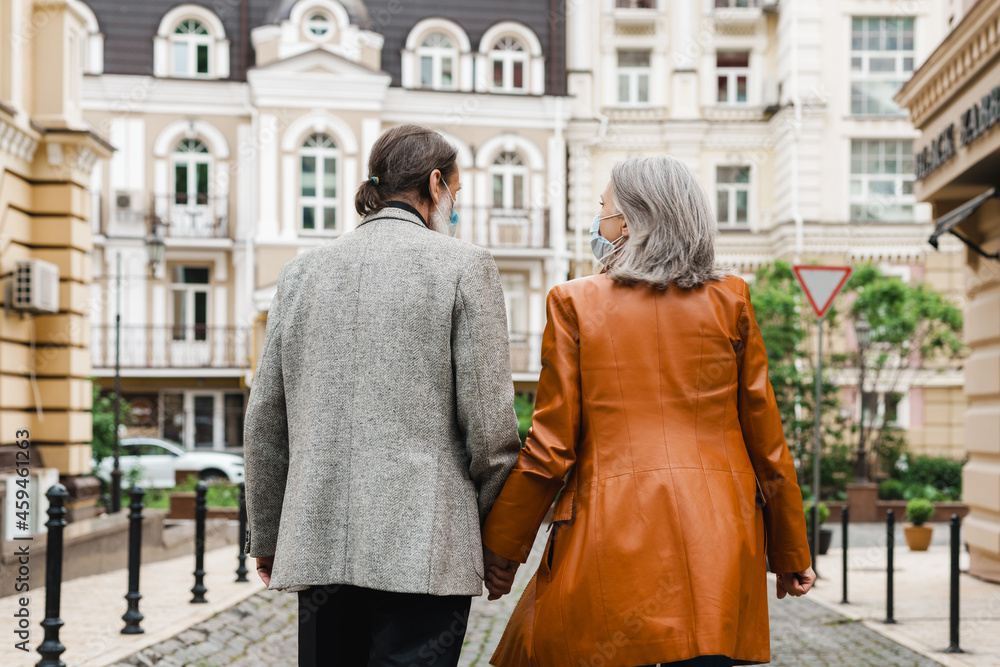 White senior couple in face masks holding hands while walking on street