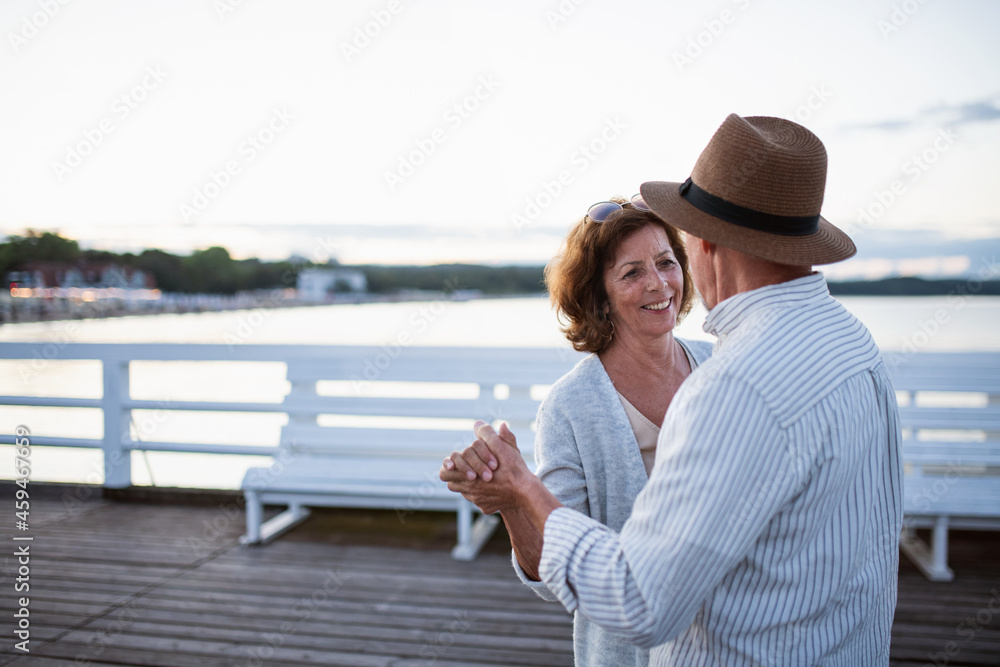 Happy senior couple dancing outdoors on pier by sea, looking at each other.