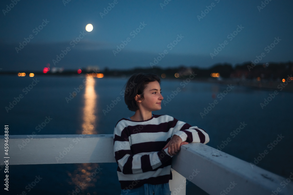 Fototapeta premium Portrait of preteen girl looking aside outdoors on pier by sea at dusk, holiday concept.