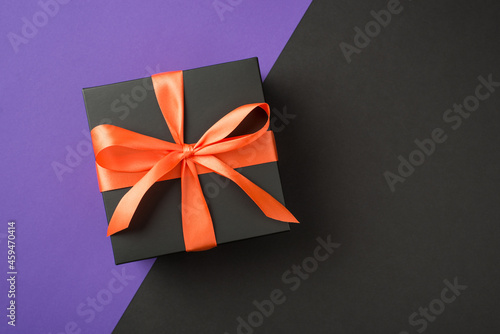Top view photo of black giftbox with orange satin ribbon bow on isolated bicolor violet and black background with empty space