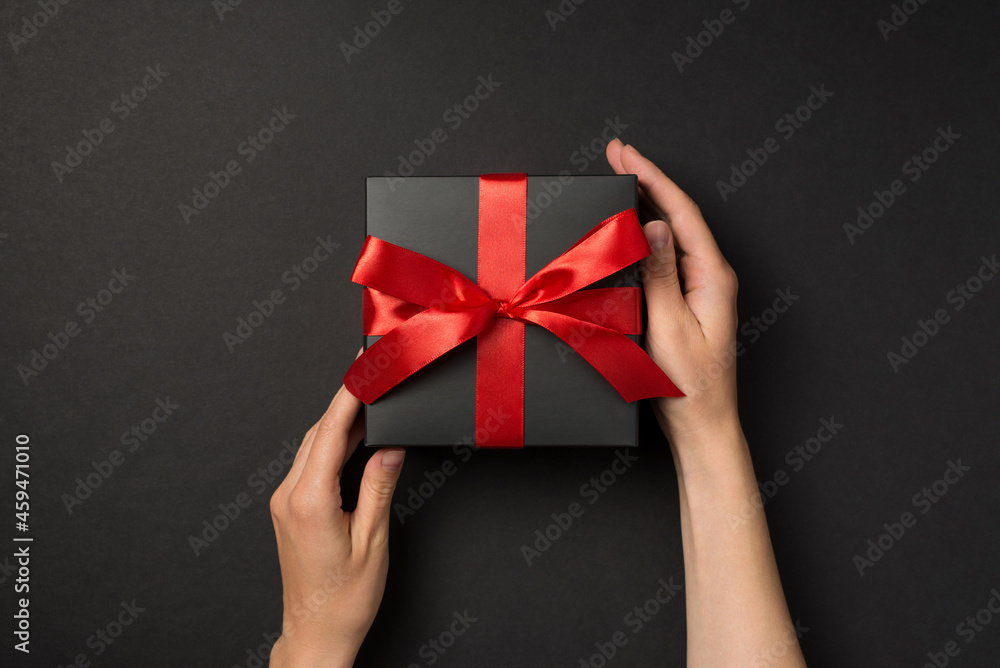 First person top view photo of woman hands touching stylish black giftbox with red satin ribbon bow on isolated black background First person top view photo of woman hands touching stylish black giftbox with red satin ribbon bow on isolated black background