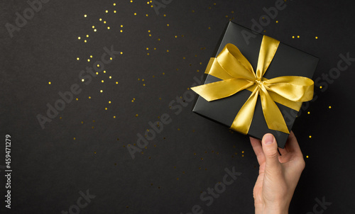 First person top view photo of hand holding black giftbox with yellow satin ribbon bow over shiny golden sequins on isolated black background with copyspace