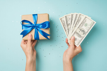 First person top view photo of hands holding fan of money and craft paper giftbox with blue ribbon bow over shiny sequins on isolated pastel blue background
