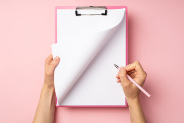 First person top view photo of female hands with pink clipboard holding pen and turning page on isolated pastel pink background with copyspace