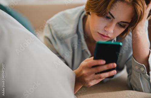 Portrait of young woman using smartphone, lying on sofa at home, social networks concept.