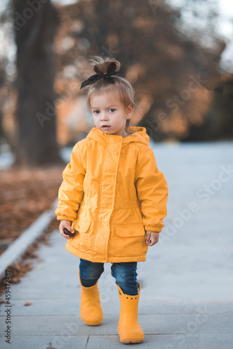 Pretty funny kid girl 2-3 year old wear yellow bright raincoat, rubber boots walk in park over fallen leaves outdoors. Autumn season. Happy child over nature background at city street. © morrowlight