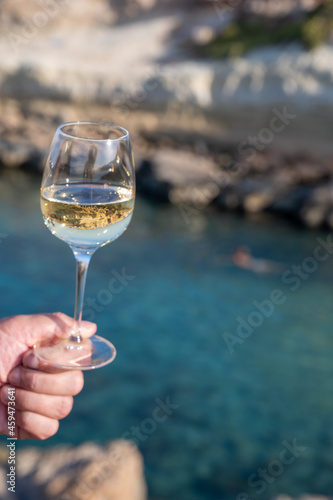 Man's hand holding glass of white dry white wine with view on rocks and blue sea bay water near Protaras touristic town on Cyprus