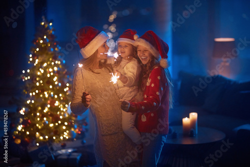 Beautiful caucasian mother hugging her two pretty daughters during new year celebration at home. Happy family standing with sparklers in hands near shiny christmas tree. Festive mood.