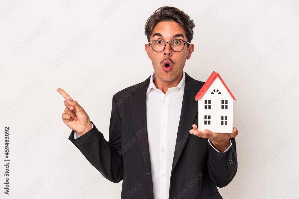 Young mixed race business man holding a toy house isolated on white background pointing to the side Young mixed race business man holding a toy house isolated on white background pointing to the side