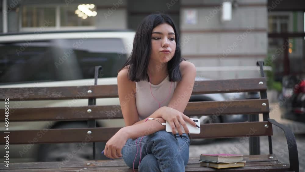 Front view of sad thoughtful tired Caucasian teenage girl listening to playlist in earphones sitting on city street on bench. Portrait of stressed adolescent teenager listening music thinking Front view of sad thoughtful tired Caucasian teenage girl listening to playlist in earphones sitting on city street on bench. Portrait of stressed adolescent teenager listening music thinking