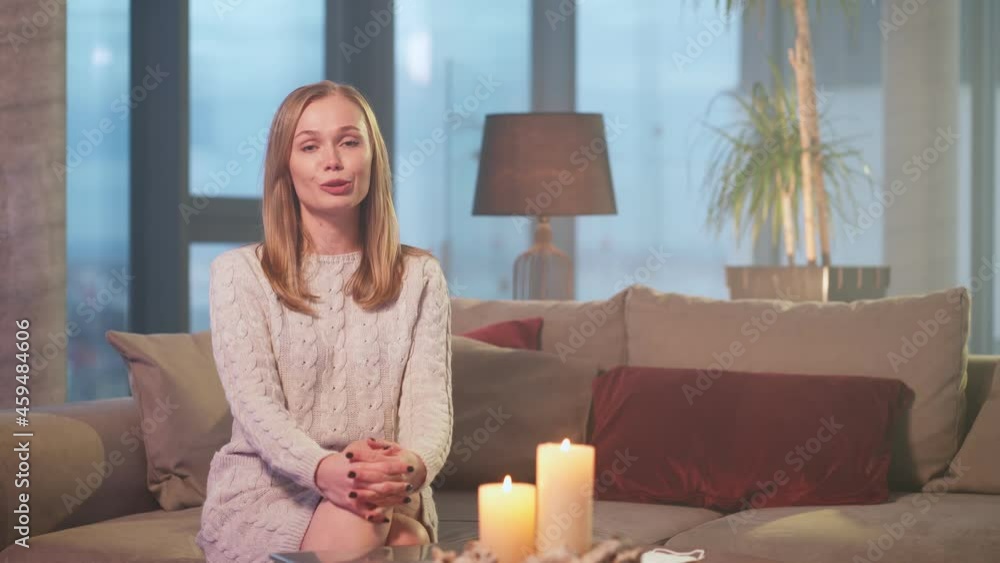 Attractive young woman with blond hair sitting on couch, smiling, waving hand and talking on camera. Concept of job interview, coaching or blogging. Attractive young woman with blond hair sitting on couch, smiling, waving hand and talking on camera. Concept of job interview, coaching or blogging.