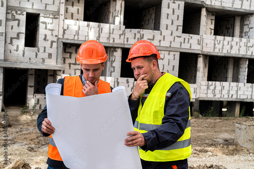on a construction site, one worker in uniform shows another worker in uniform a plan of the house on a construction site, one worker in uniform shows another worker in uniform a plan of the house