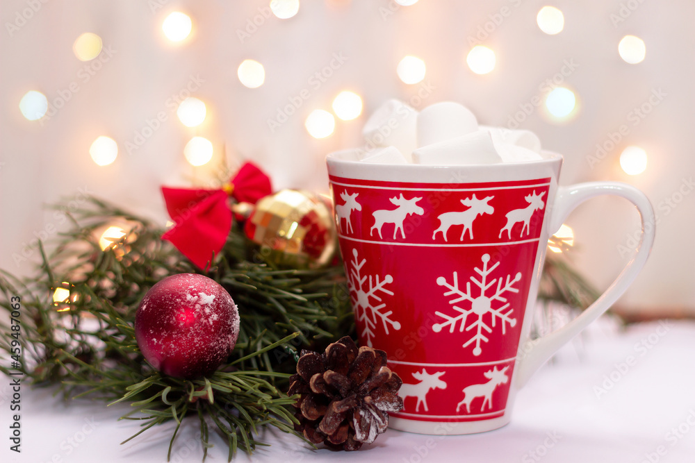 A red mug with a deer, snowflakes pattern with beverage and marshmallow near a branch of a Christmas tree, toy balls and cone on a background of blurred bokeh lights of garland. New Year's decorations A red mug with a deer, snowflakes pattern with beverage and marshmallow near a branch of a Christmas tree, toy balls and cone on a background of blurred bokeh lights of garland. New Year's decorations