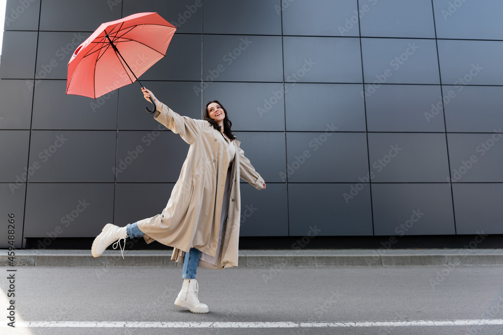 happy woman in beige coat and white boots posing with red umbrella in raised hand - obrazy, fototapety, plakaty 