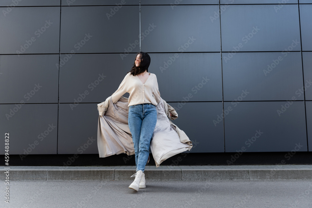 cheerful woman in jeans and white boots posing with raincoat near grey wall cheerful woman in jeans and white boots posing with raincoat near grey wall