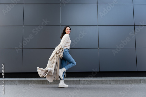 happy woman in jeans and white boots smiling at camera while running along grey wall © LIGHTFIELD STUDIOS