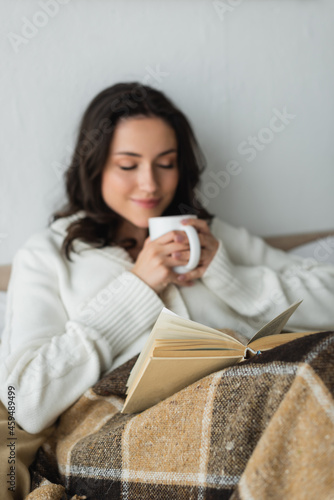 blurred woman with closed eyes holding cup of tea while resting in bed with book