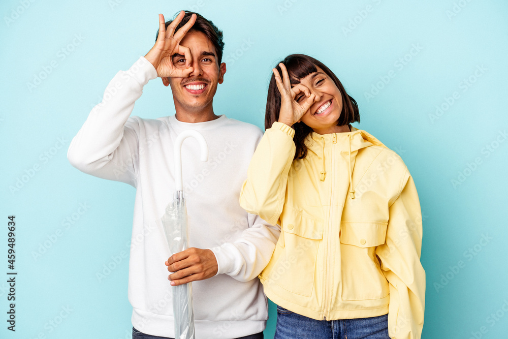 Fototapeta premium Young mixed race couple holding umbrella isolated on blue background excited keeping ok gesture on eye.