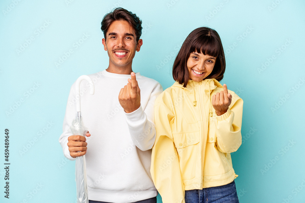 Young mixed race couple holding umbrella isolated on blue background pointing with finger at you as if inviting come closer. Young mixed race couple holding umbrella isolated on blue background pointing with finger at you as if inviting come closer.