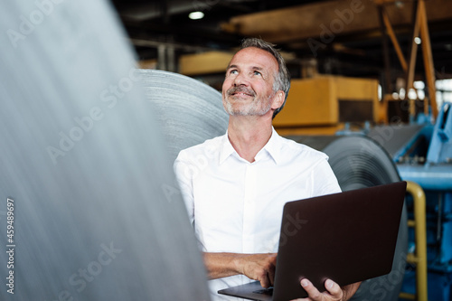 Smiling male inspector with laptop checking metal sheet roll in steel mill photo