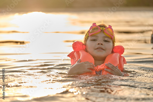 A little girl bathes in the water in the sunset light. Child playing on the beach during summer vacation. The child swims in a life jacket and goggles. Safety on the water.