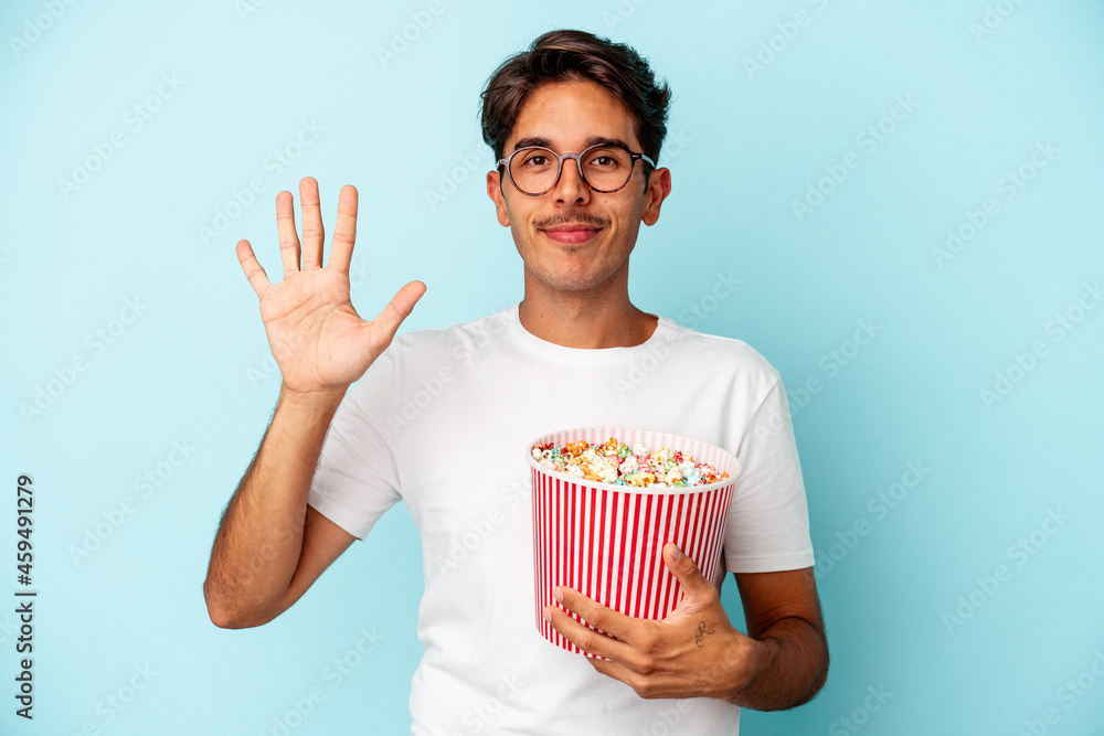 Young mixed race man eating popcorns isolated on blue background smiling cheerful showing number five with fingers. Young mixed race man eating popcorns isolated on blue background smiling cheerful showing number five with fingers.