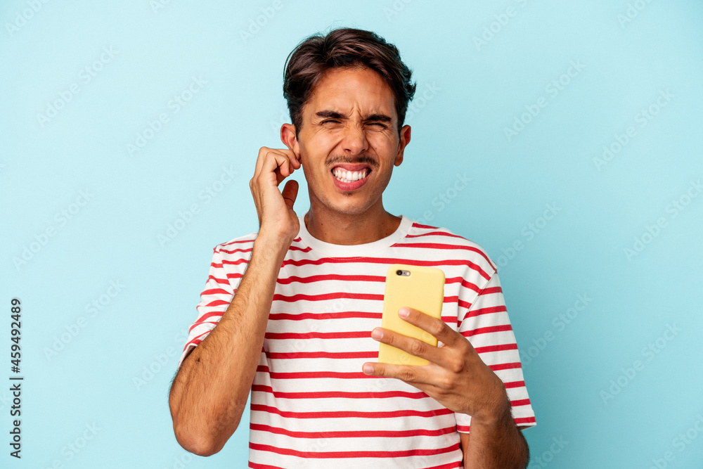 Young mixed race man holding mobile phone isolated on blue background covering ears with hands. Young mixed race man holding mobile phone isolated on blue background covering ears with hands.