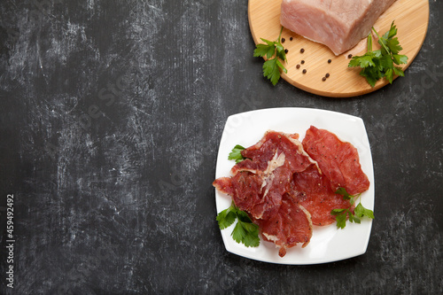 Pieces of jerky on a white porcelain plate and raw meat on a cutting board on a black background. Copy spaes.