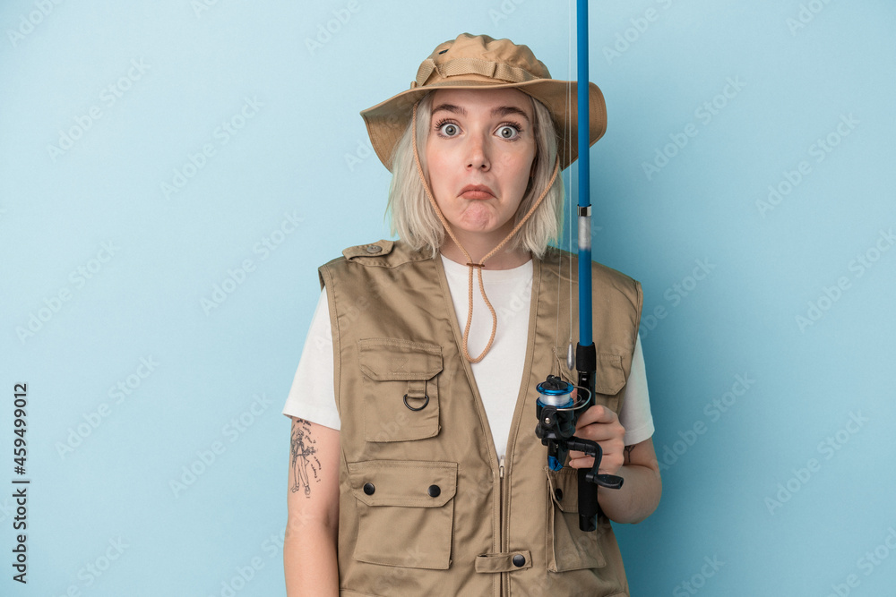 Young caucasian fisherwoman holding a rod isolated on blue background shrugs shoulders and open eyes confused. Young caucasian fisherwoman holding a rod isolated on blue background shrugs shoulders and open eyes confused.