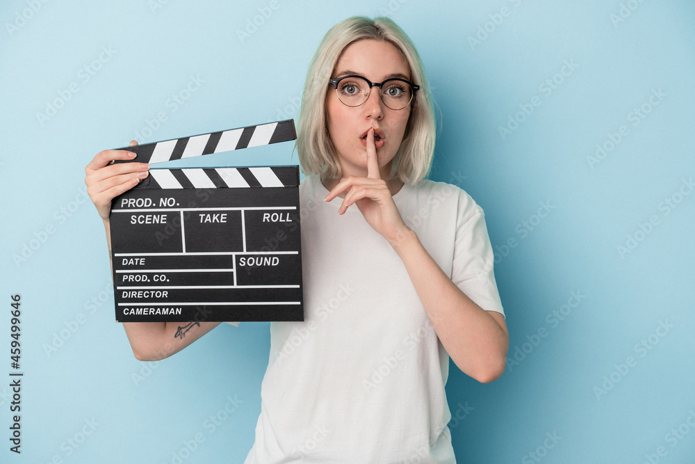 Young caucasian woman holding a clapperboard isolated on blue background keeping a secret or asking for silence. Young caucasian woman holding a clapperboard isolated on blue background keeping a secret or asking for silence.