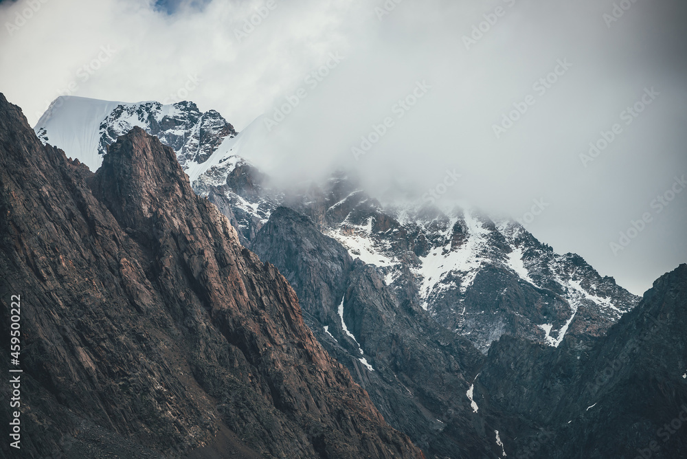 Awesome mountain landscape with snowy mountains in low clouds. Rocky top and snowy top. Atmospheric view to great rocky mountains in cloudy sky. Gloomy scenery with high mountains in overcast weather.