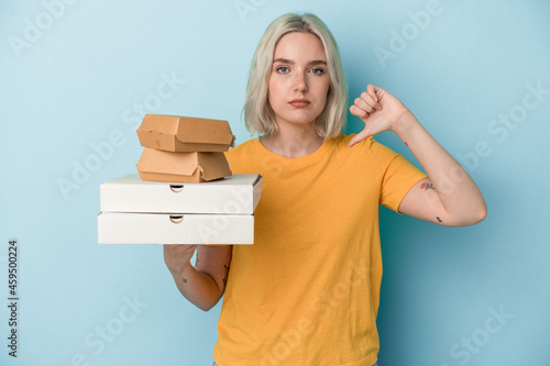 Young caucasian woman holding pizzas and burgers isolated on blue background showing a dislike gesture, thumbs down. Disagreement concept.