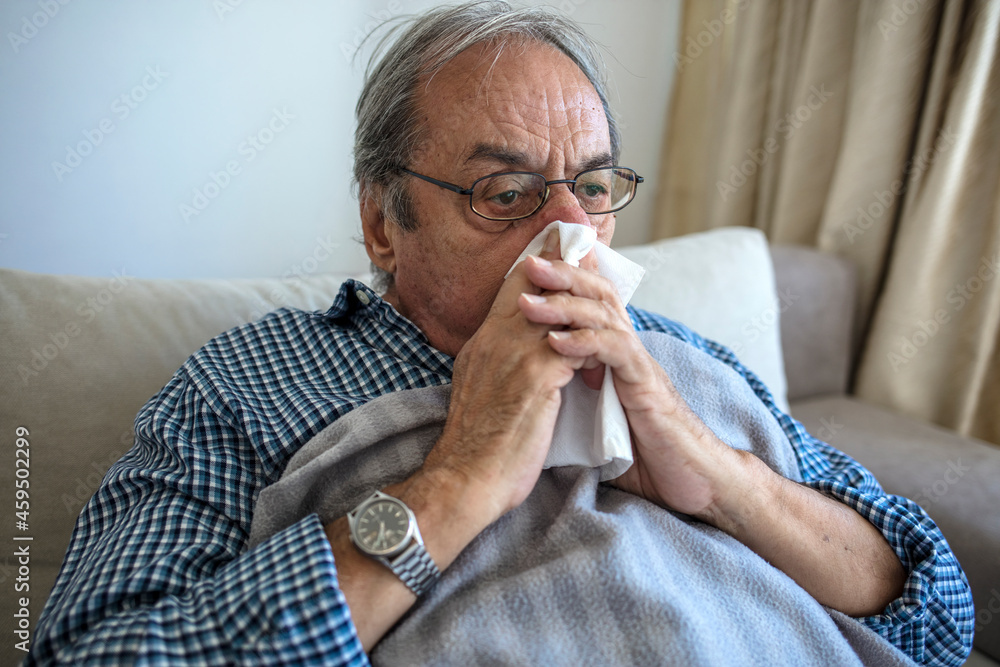 Elderly unwell man lying on couch at home and blowing nose. Sick old man with cold lying in bed and blow nose. Man feeling cold, lying in the bed and blowing his nose at home during the day. Elderly unwell man lying on couch at home and blowing nose. Sick old man with cold lying in bed and blow nose. Man feeling cold, lying in the bed and blowing his nose at home during the day.