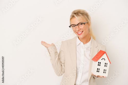 Young real estate agent woman holding a home model isolated on white background showing a copy space on a palm and holding another hand on waist.