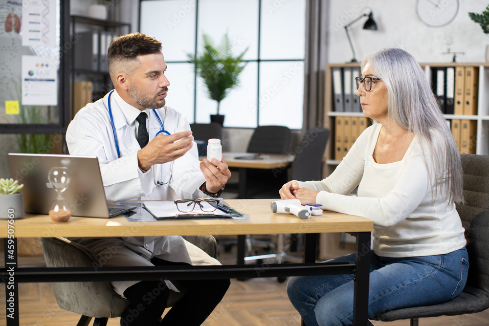 Fototapeta premium Qualified male doctor in uniform sitting at workplace with aged woman and providing medical consultation, talking about new medications. Concept of people, medicine and health care.
