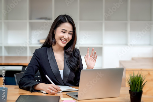 Image of a beautiful Asian businesswoman waving greetings online through laptop holding pen and notebook at the office. © amnaj