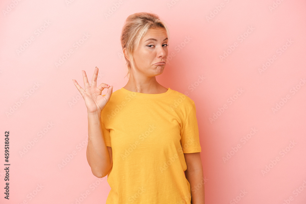 Obraz premium Young Russian woman isolated on pink background winks an eye and holds an okay gesture with hand.