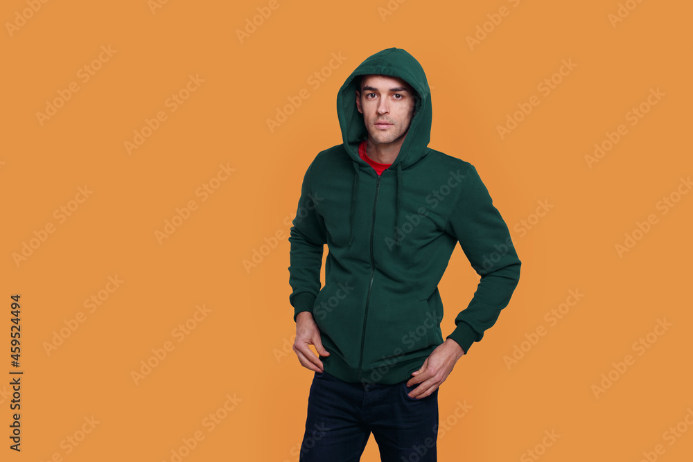 Portrait of a young man in green jumper and jeans, with a hood on his head, isolated orange background. Portrait of a young man in green jumper and jeans, with a hood on his head, isolated orange background.