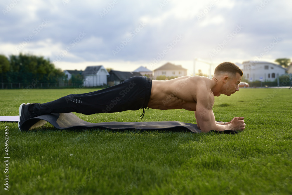 Fototapeta premium Muscular man with naked torso standing in plank position on yoga mat outdoors. Caucasian bodybuilder doing exercises for abs on grass of stadium. 