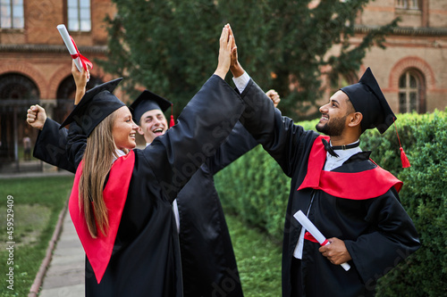 Three happy international graduate friends greeting in university campus in graduation robes with diploma